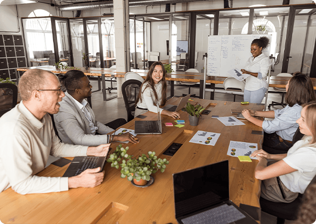 Group of coworkers standing together in an office, listening to a man with glasses speaking during a workplace gathering.
